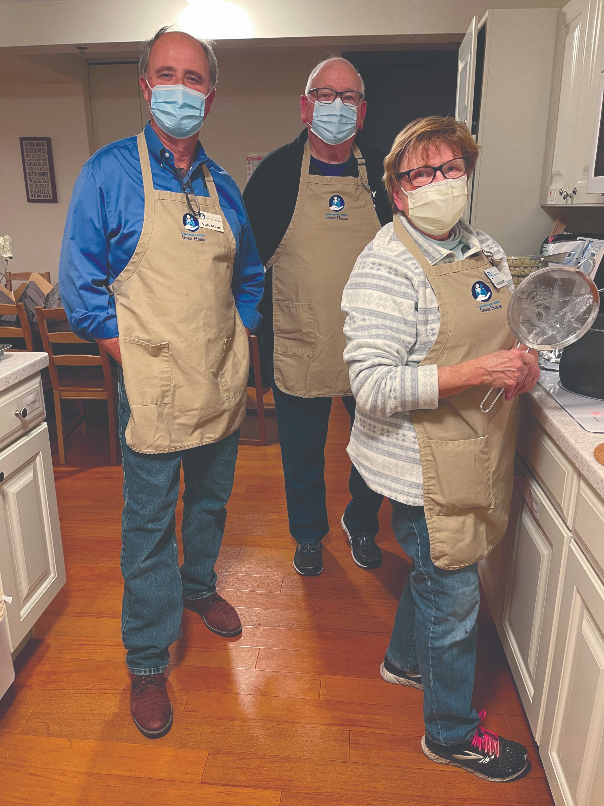 Three voluneers stand in a kitchen wearing masks and beige aprons embroidered with the Guest House logo.