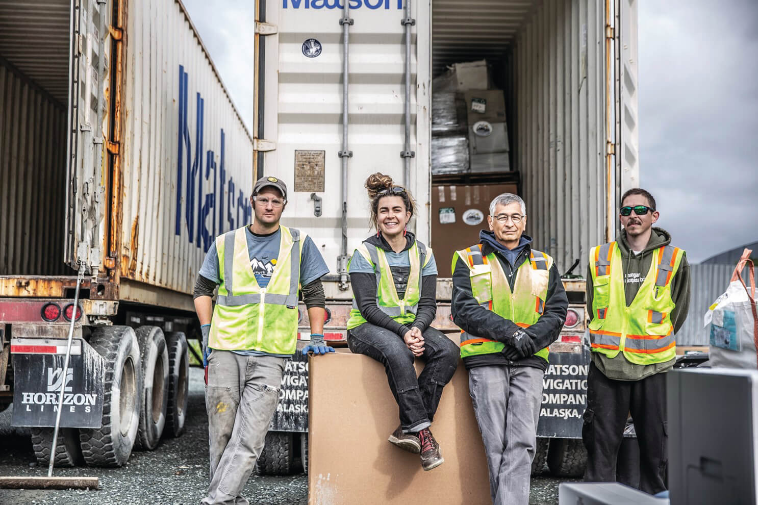 Four Backhaul stafff and volunteers wearing yellow safety vests pose in front of a Matson container loaded with e-waste.