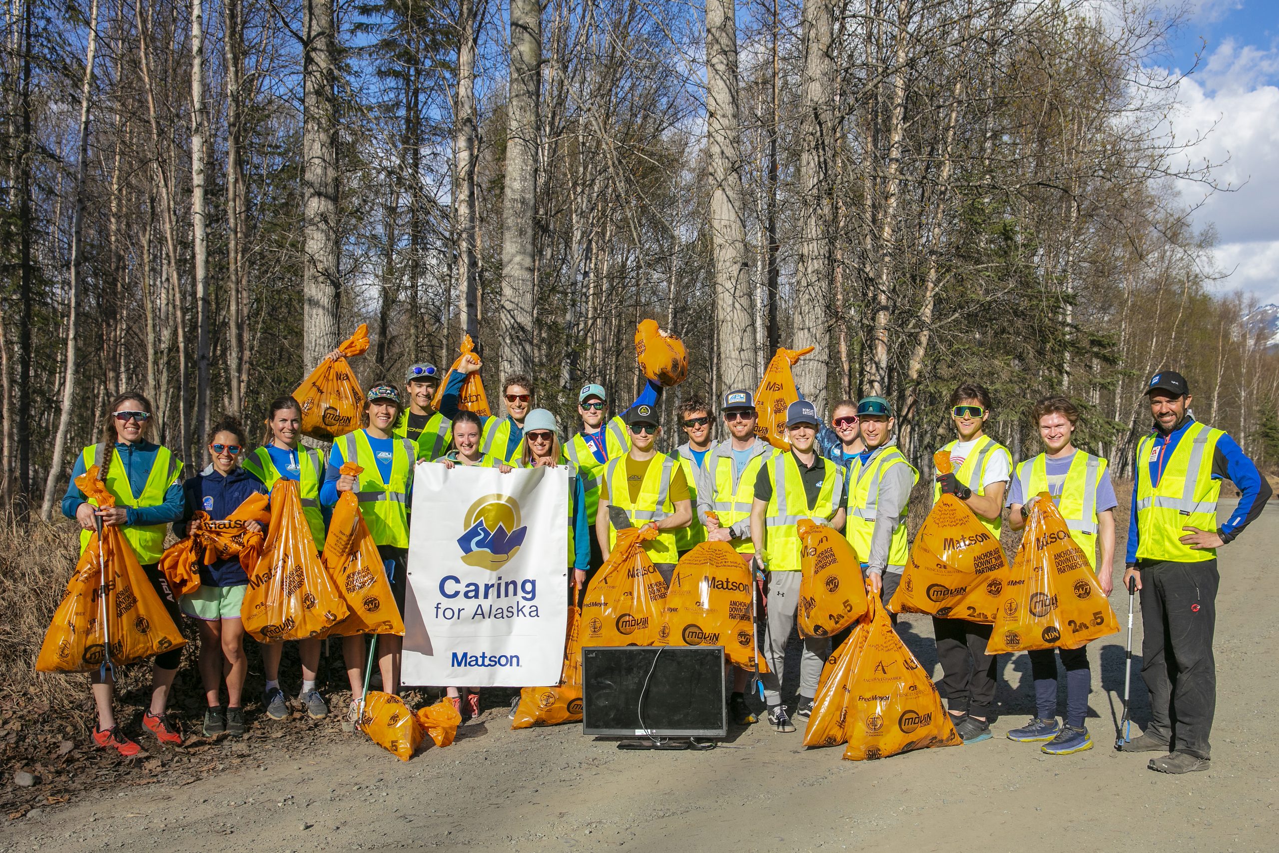 Volunteers wearing yellow safety vests pose for a group picture with their full orange trash bags and Caring For Alaska banner.