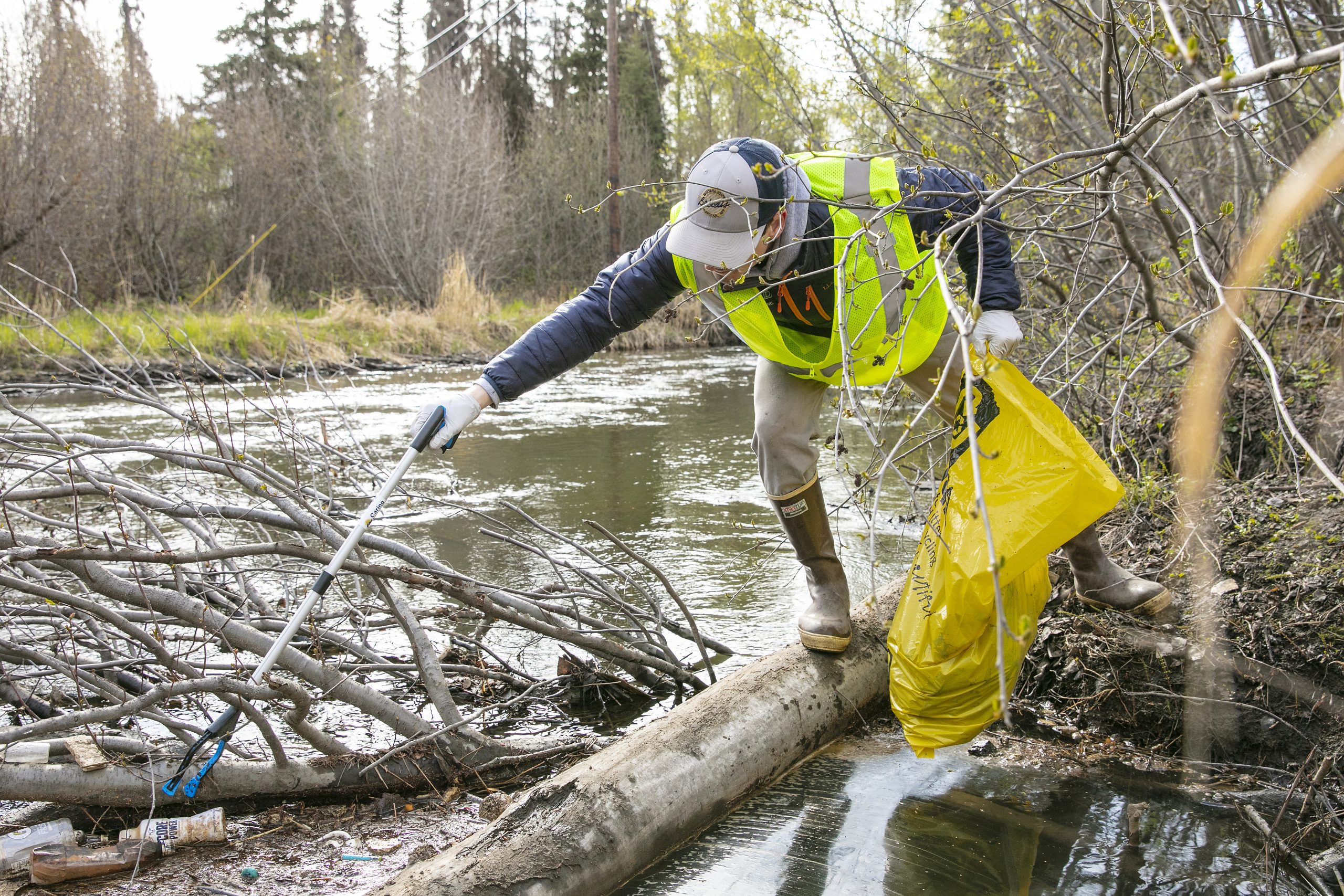 A volunteer wearing rubber boots and yellow safety vest holds a yellow trash bag while reaching with a grabber to pick up trash in Campbell Creek.