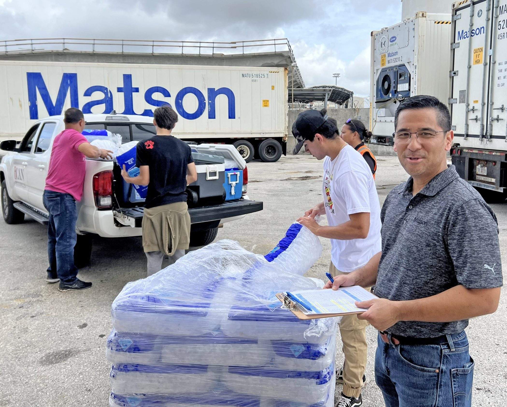 Matson employees move bags of ice from a pallet into ice chests with Matson reefer and containers in the background.