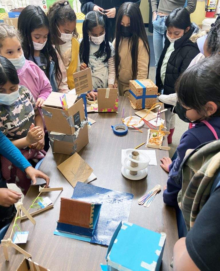 Girls gather shoulder to shoulder around a table to view a variety of creations made of tape, cardboard, paper, duct tape, paper, popsicle sticks, pipe cleaners, and more.