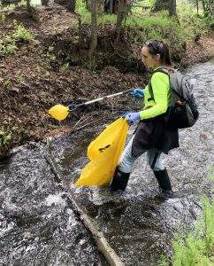 A woman wearing rubber boots, a gray backpack, work gloves, and a bright green shirt stands in a creek with a grabber and yellow plastic bag to pick up trash.