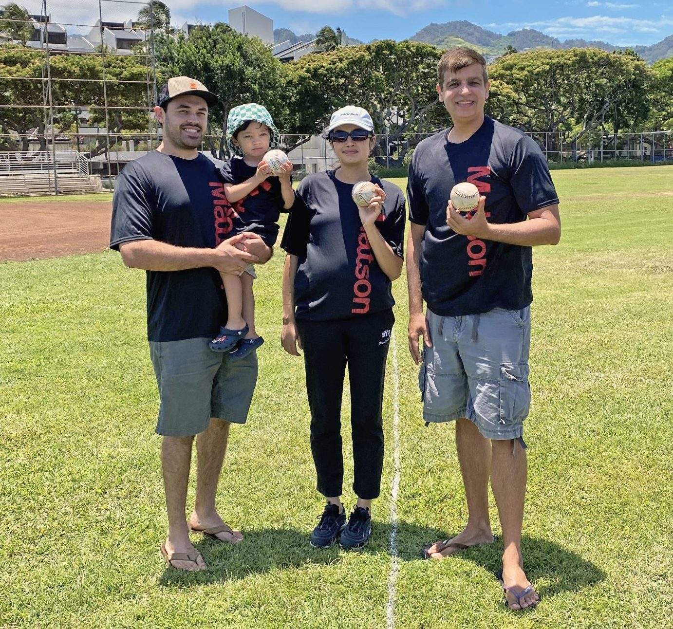 A family of three adults and one small child wear black t-shirts with the red Matson logo and hold white softballs in the outfield.