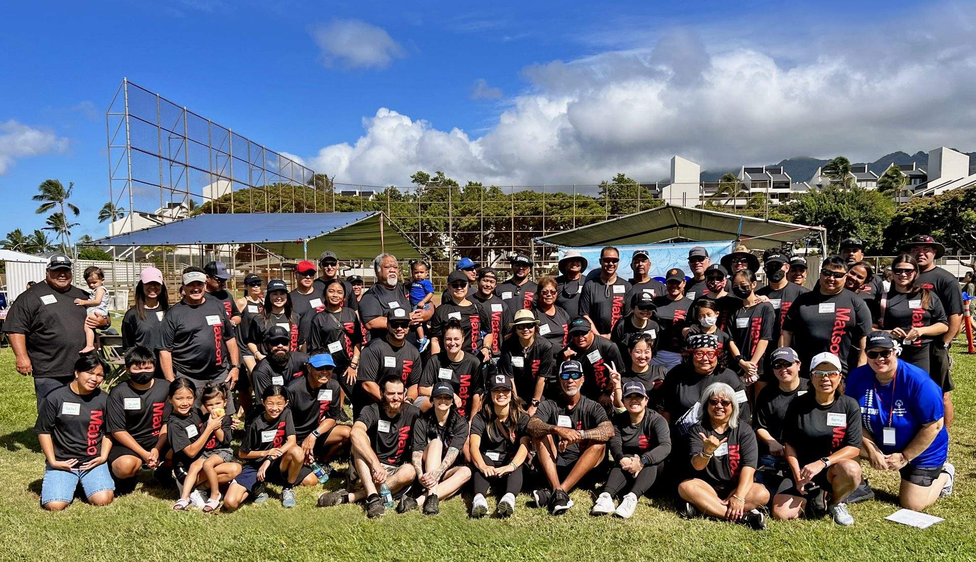 Matson volunteers dressed in black t-shirts with a red Matson logo pose for a group picture with the softball field backstop in the background.