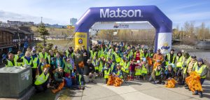 Matson volunteers wearing yellow vests and holding orange trash bags pose in front of a Matson logoed blue arch with downtown Anchorage in the background.